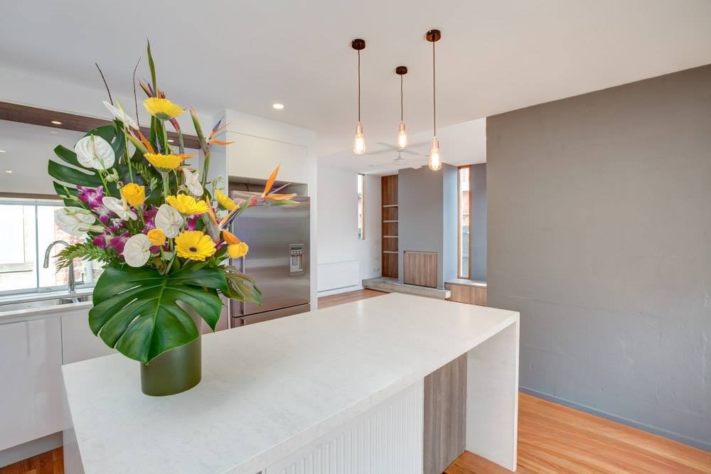 A modern kitchen interior featuring a white countertop, a tall vase with colorful flowers, and pendant lighting overhead.