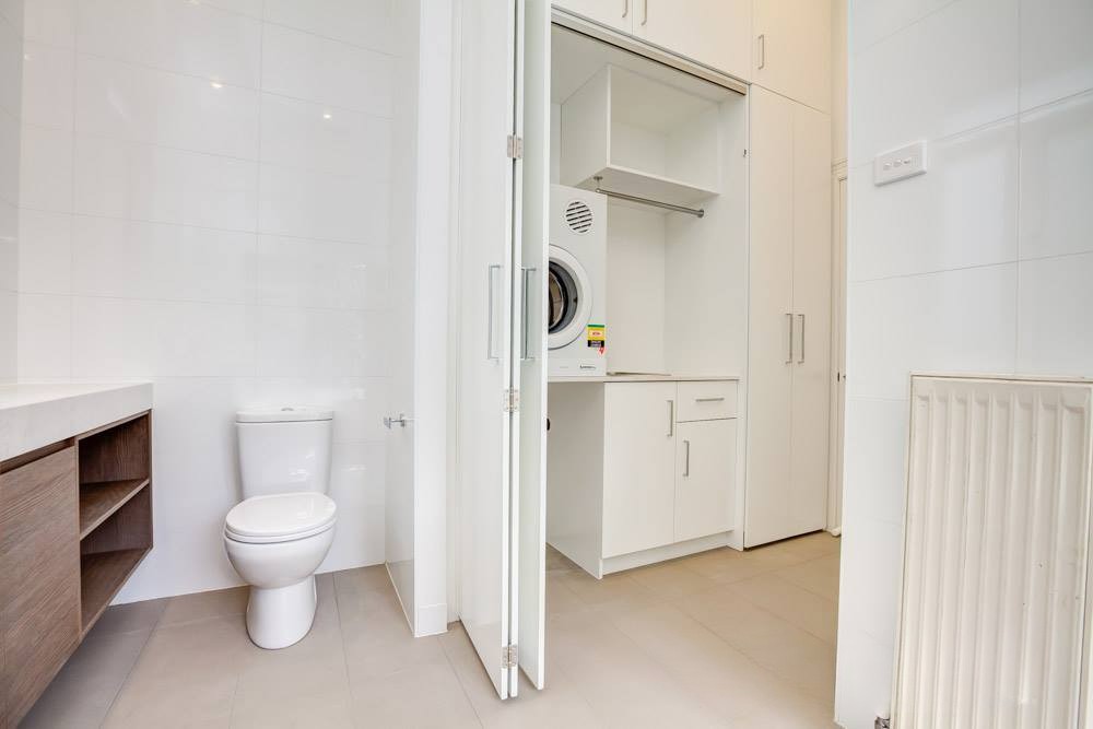 A modern bathroom featuring a white toilet, a wooden vanity, and a laundry area with a washing machine and storage cabinets.