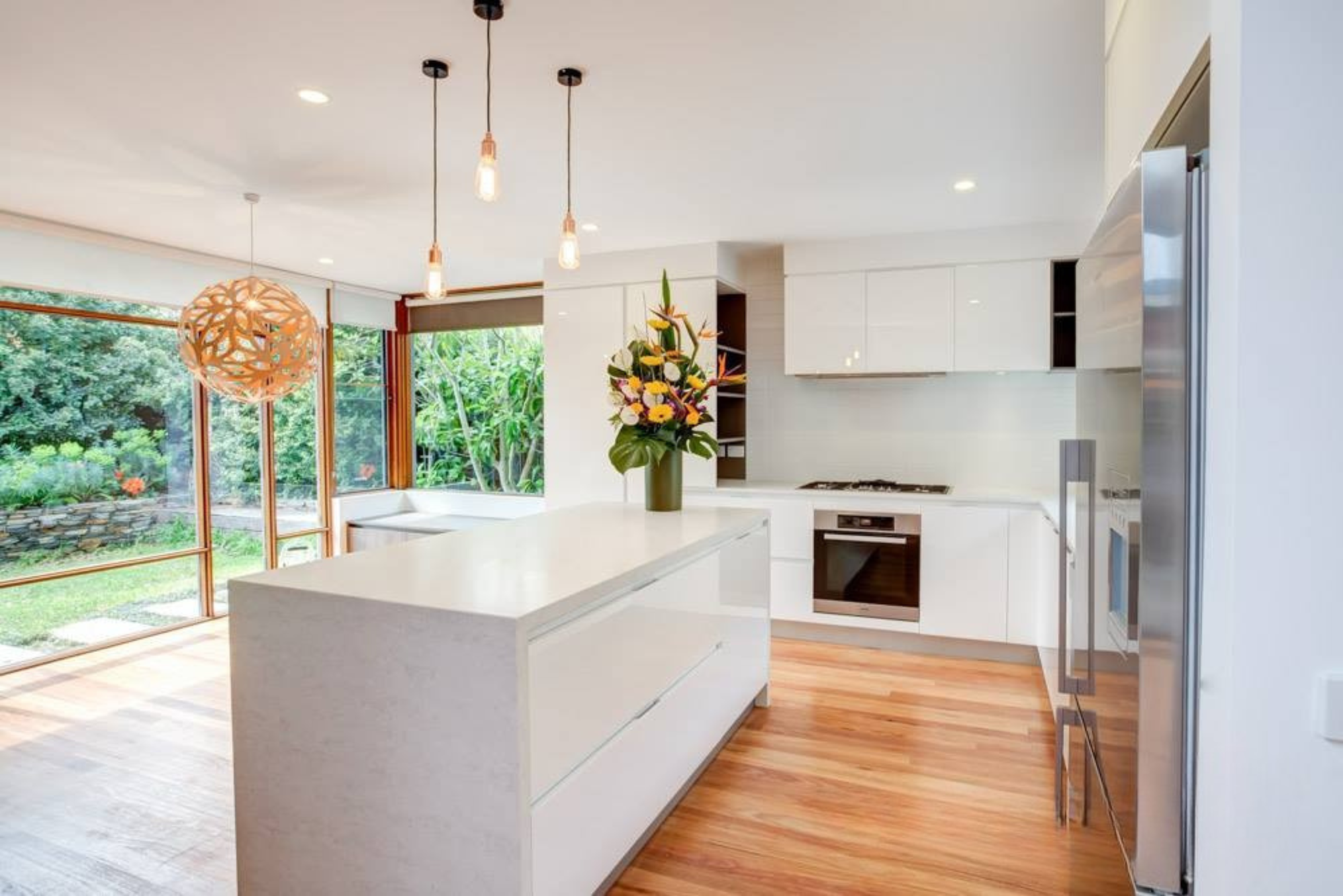 A modern kitchen featuring white cabinets, a large island with a sleek countertop, and pendant lighting. A bouquet of flowers is displayed on the island, and large windows provide a view of green landscaping outside.