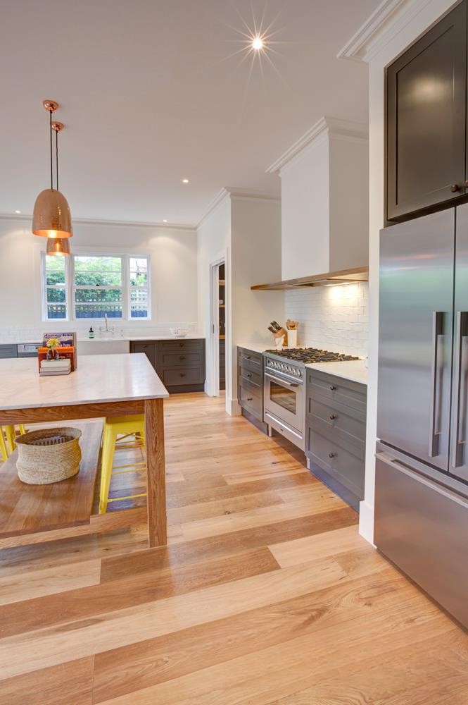 Modern kitchen with a large island featuring a wooden table and yellow chairs, stainless steel appliances, gray cabinets, and wooden flooring.