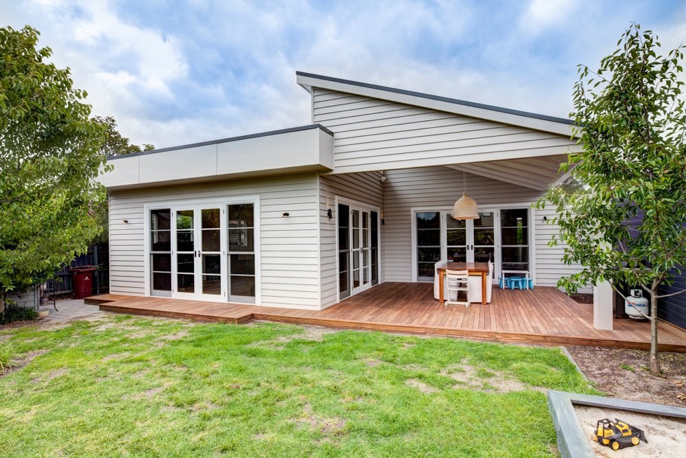 Exterior view of a modern house with a wooden deck, large glass doors, and surrounding greenery.