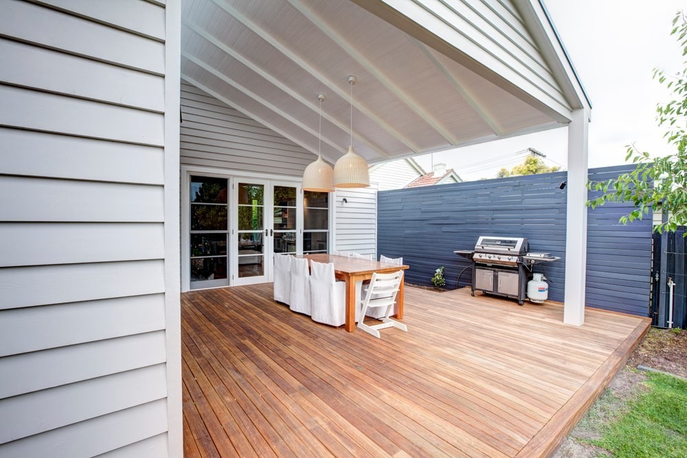 A wooden deck featuring a dining table with white chairs, two pendant lights, and a barbecue grill, surrounded by a blue wooden fence.