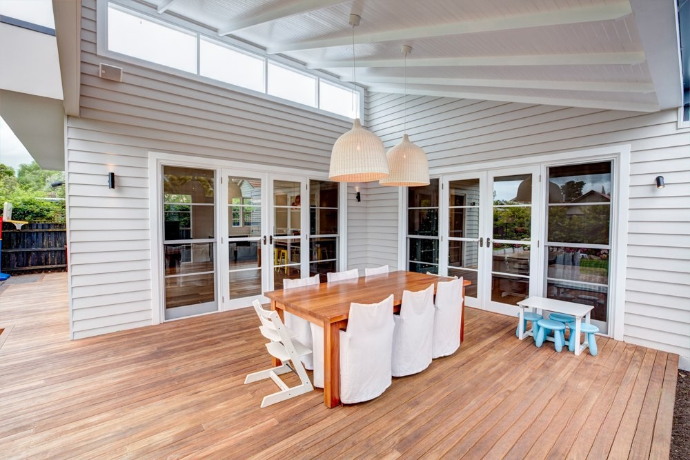 Stylish outdoor dining area with a wooden table and white chairs beneath two pendant lights, surrounded by large windows in a modern home setting.