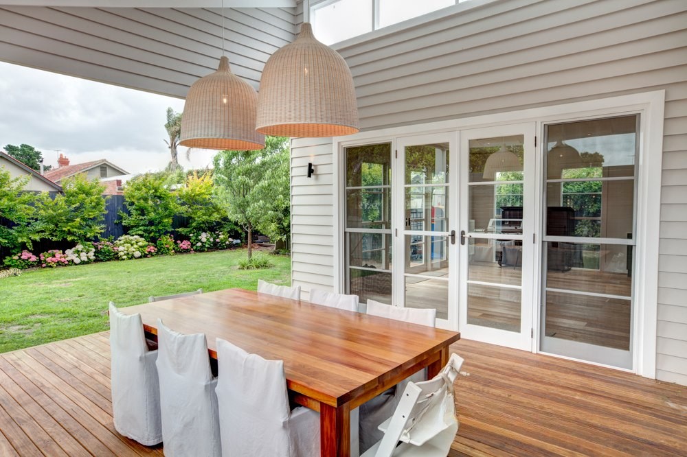 An outdoor dining area featuring a wooden table with white-covered chairs, large woven pendant lights, and sliding glass doors leading into a light-filled interior. A lush garden with green grass and flowering shrubs is visible in the background.
