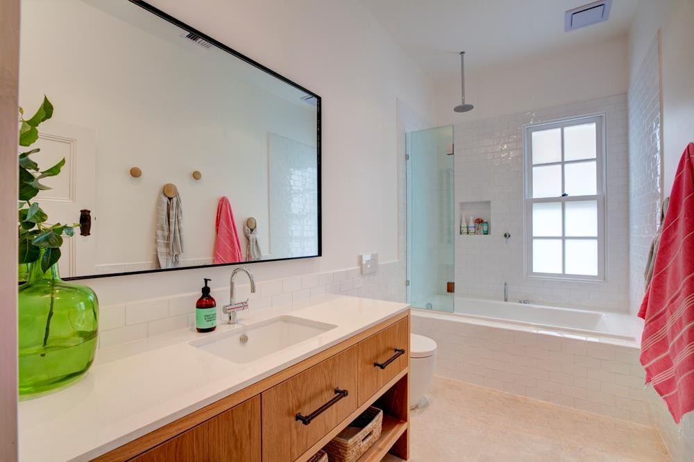 Modern bathroom featuring a large mirror, wooden vanity, and a green vase with plants. The space includes a bathtub, shower area with glass enclosure, and towels hanging on hooks.