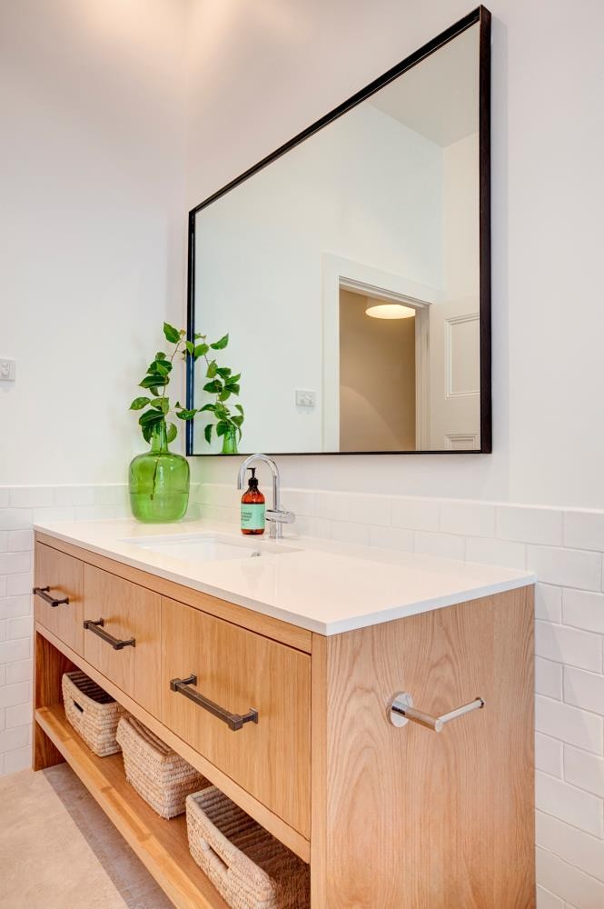 A modern bathroom with a wooden vanity, white countertop, and a large mirror. The vanity features storage baskets and a stylish green plant beside a bottle of soap.