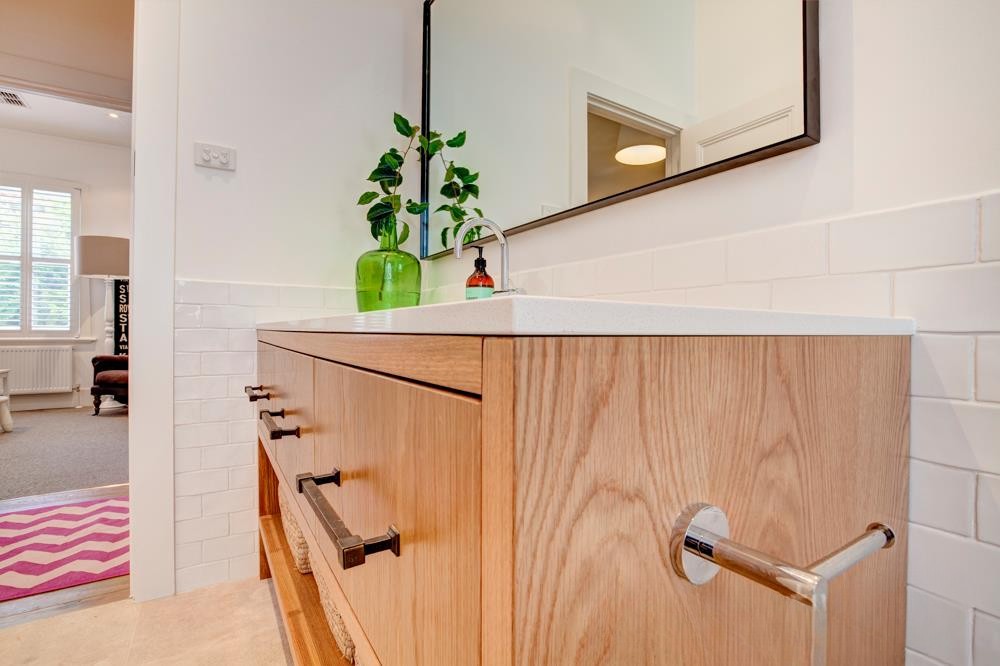 A modern bathroom vanity with a wooden finish, white countertop, and a sleek faucet. A green glass vase with plants is placed on the countertop, alongside a small brown bottle. The background features white tiles and a mirror.