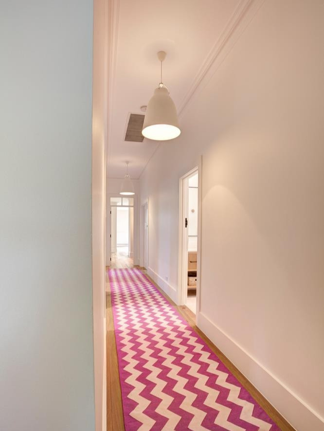 A well-lit hallway featuring a zigzag patterned rug in pink and white. The walls are light-colored, and there are simple pendant lights hanging from the ceiling.