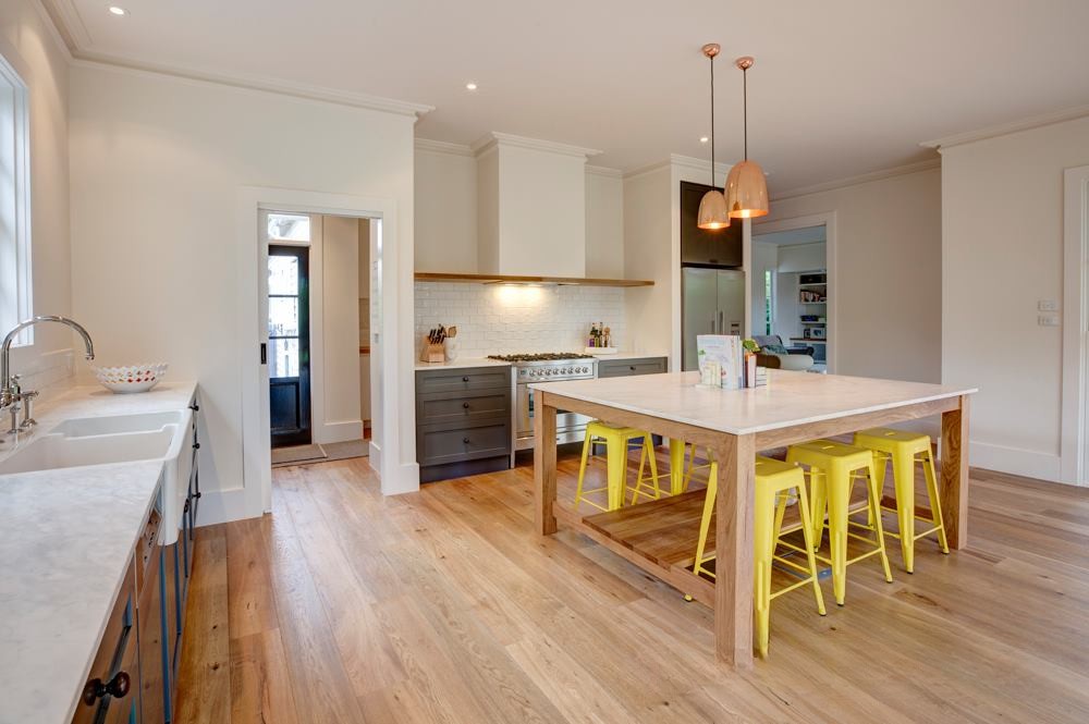 A modern kitchen featuring a large island with a wooden base and white countertop, complemented by bright yellow stools. The kitchen includes gray cabinets, a farmhouse sink, and pendant lighting.