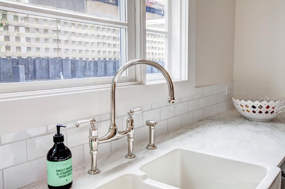 A modern kitchen sink area featuring a polished metal faucet, a soap dispenser with a green label, and a decorative bowl. Bright natural light streams through the windows, illuminating the marble countertop and white subway tile backsplash.