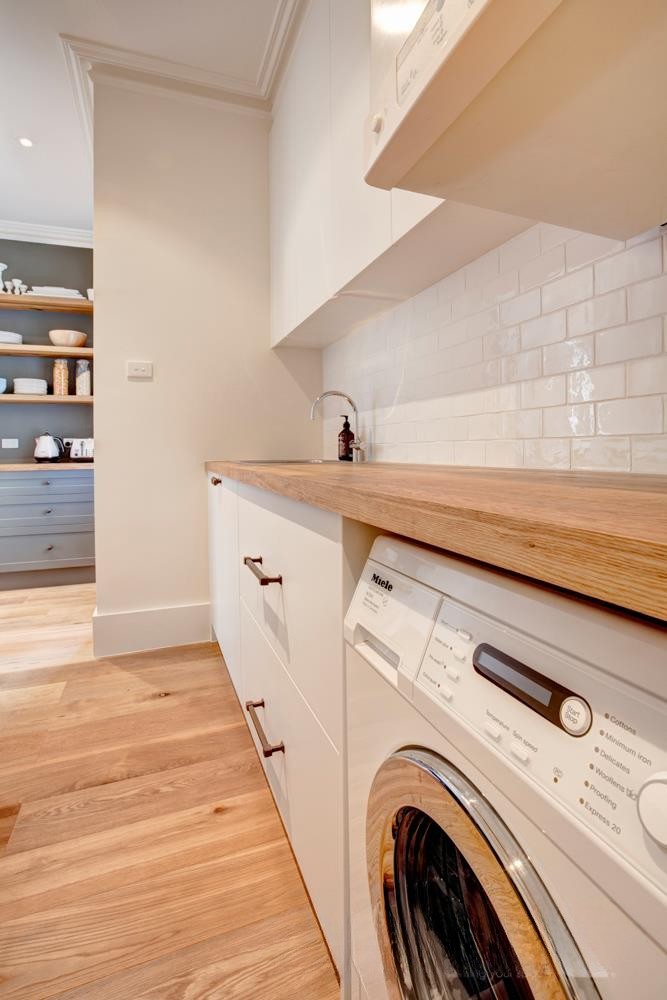 A modern laundry room featuring a Miele washing machine, wooden countertop, white cabinetry, and a neatly arranged shelf in the background.