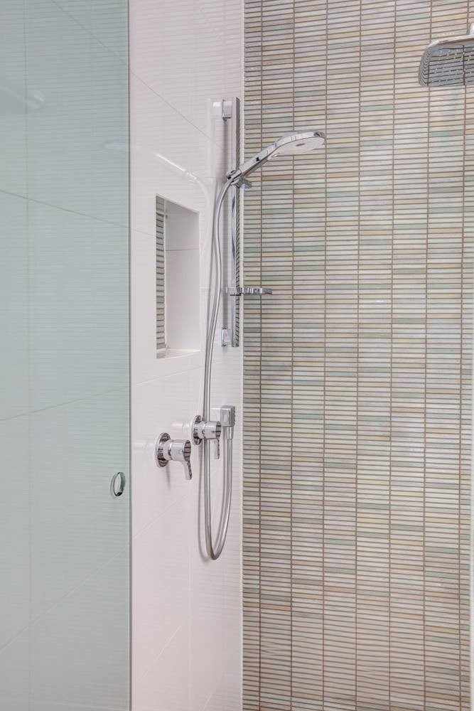Modern shower setup featuring a wall-mounted showerhead, handheld shower, and glass partition with textured tile background.
