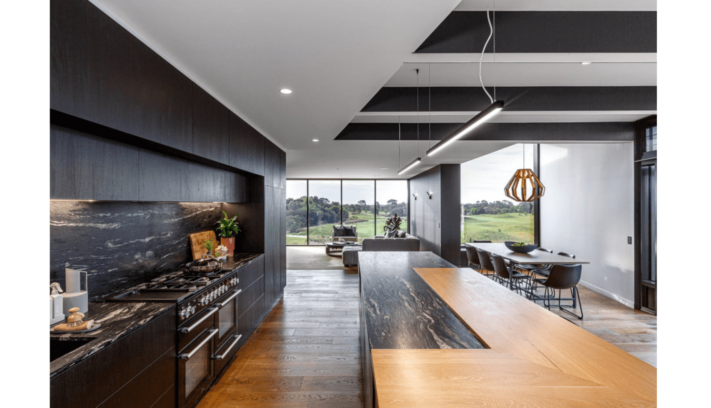Modern kitchen featuring dark cabinetry, a marble backsplash, and a large wooden island, with a dining area and scenic outdoor view in the background.