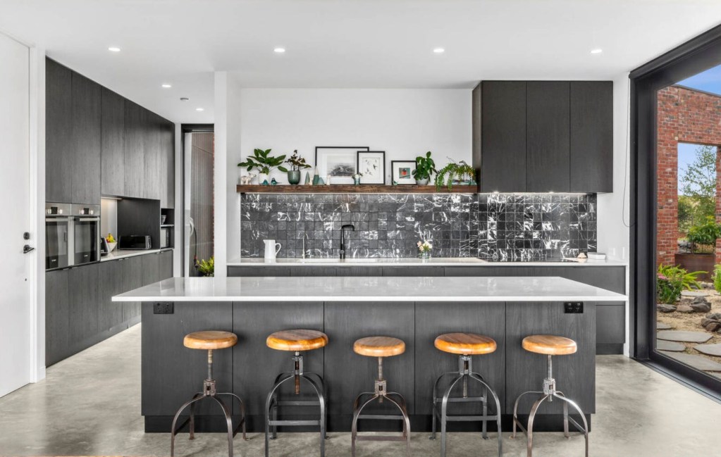 Modern kitchen featuring a large island with three wooden stools, sleek dark cabinetry, a marble backsplash, and large windows showcasing a garden view.